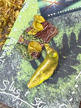 Load image into Gallery viewer, Handmade forest floor pin featuring a golden-yellow banana slug climbing a miniature mossy stump with tiny sculpted chanterelles. Polymer clay, hand-painted, pin back. Displayed on a dark green card with pressed fern design and hand-lettered "Slug & Stump" in silver ink.by the woodland wandolier