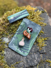 Load image into Gallery viewer, Hand sculpted peachy bronze snail shell with mushroom colony on its back, with a tiny round bottle of salt dangling below it via brass findings.made by The Woodland Wandolier , sitting on a mossy grey stone with business card and packaging.