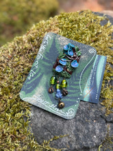 Load image into Gallery viewer, Hand sculpted polymer forest themed brooch, by The Woodland Wandolier.
Detailed with vines, leaves, and blue cup fungi, green foil beads, tiny lapis frog, bell, and miniature animal foot print beads dangling below it. photographed on a grey stone with moss on it.