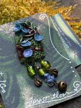 Load image into Gallery viewer, Hand sculpted polymer forest themed brooch, by The Woodland Wandolier.
Detailed with vines, leaves, and blue cup fungi, green foil beads, tiny lapis frog, bell, and miniature animal foot print beads dangling below it. photographed on a grey stone with moss on it.