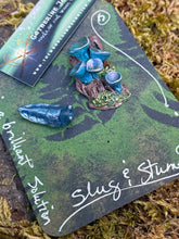 Load image into Gallery viewer, Handmade forest floor themed pin featuring a fantasy slug with love darts perched on a miniature stump with iridescent blue elf cup mushrooms. Polymer clay, hand-painted, pin back. Displayed on a dark green card with pressed fern design and hand-lettered "Slug & Stump" in silver ink. Handsculpted by The Woodland Wandolier