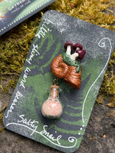 Load image into Gallery viewer, Hand sculpted peachy bronze snail shell with mushroom colony on its back, with a tiny round bottle of salt dangling below it via brass findings.made by The Woodland Wandolier , sitting on a mossy grey stone with business card and packaging.
