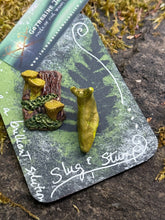 Load image into Gallery viewer, Handmade forest floor pin featuring a golden-yellow banana slug climbing a miniature mossy stump with tiny sculpted chanterelles. Polymer clay, hand-painted, pin back. Displayed on a dark green card with pressed fern design and hand-lettered "Slug &amp; Stump" in silver ink.by the woodland wandolier 
