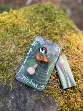 Load image into Gallery viewer, Hand sculpted peachy bronze snail shell with mushroom colony on its back, with a tiny round bottle of salt dangling below it via brass findings.made by The Woodland Wandolier , sitting on a mossy grey stone with business card and packaging.
