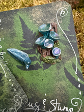 Load image into Gallery viewer, Handmade forest floor themed  pin featuring a fantasy slug with love darts perched on a miniature stump with iridescent blue elf cup mushrooms. Polymer clay, hand-painted, pin back. Displayed on a dark green card with pressed fern design and hand-lettered "Slug &amp; Stump" in silver ink. Handsculpted by The Woodland Wandolier 

