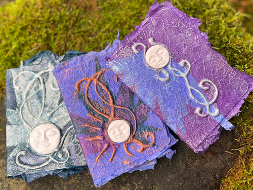 a trio of palm sized handmade journals, all in shades of purple and lavender, with moon faces and tendril designs. 