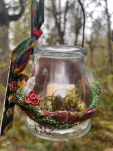 Load image into Gallery viewer, small glass candle holder that has forest style decorations all around it, hanging from a mossy branch 
