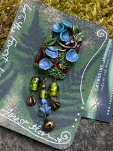 Load image into Gallery viewer, Hand sculpted polymer forest themed brooch, by The Woodland Wandolier. 
Detailed with vines, leaves, and blue cup fungi, green foil beads, tiny lapis frog, bell, and miniature animal foot print beads dangling below it.
