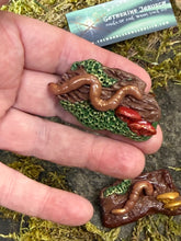 Load image into Gallery viewer, Hand-sculpted polymer clay earthworm brooch with red shelf mushrooms on a mossy stump, held in hand for scale, with business card and second brooch visible on moss

