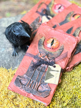 Load image into Gallery viewer, Handmade rust-colored notebook with a unique black tower design, featuring a prominent red glass eye reminiscent of Sauron from LOTR. 4 of those in a fan layout on a mossy stone, with a faux crow beside them.
