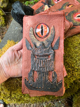 Load image into Gallery viewer, close up of Handmade rust-colored notebook with a unique black tower design, held in a hand, featuring a prominent red glass eye reminiscent of Sauron from LOTR. 4 of those in a fan layout on a mossy stone, with a faux crow beside them
