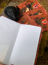Load image into Gallery viewer, Handmade rust-colored notebook with a unique black tower design, featuring a prominent red glass eye reminiscent of Sauron from LOTR. 4 of those in a fan layout, one opened to show lined paper, on a mossy stone, with a faux crow beside them
