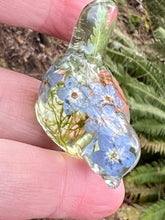 Load image into Gallery viewer, small clear resin bird figure with moss, fern, and forgetmenots inside, sitting on three fingers
